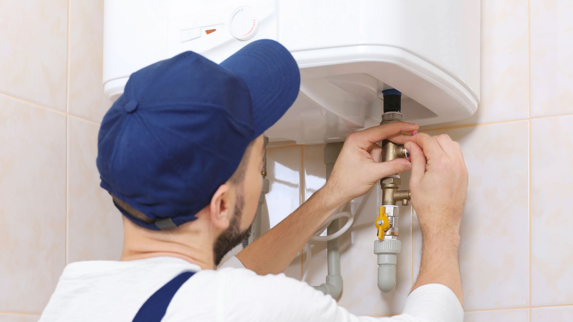 Plumber in a blue cap fitting a valve to the pipework under a wall-mounted unit