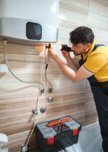 Plumber servicing a wall mounted water heater in a tiled bathroom with a toolbox on the floor